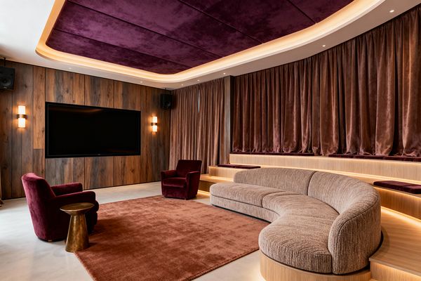 A transitional media room features a curved beige sectional on tiered wood platforms with integrated LED lighting, two burgundy velvet armchairs, and a rust rug over a light microcement floor. A walnut-paneled wall holds a large flat-screen flanked by frosted sconces and black speakers. Floor-to-ceiling mocha velvet curtains line a curved wall, and a plum velvet coffered ceiling with recessed and cove lights creates a warm, cinematic atmosphere.