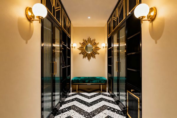 An Art Deco mudroom features symmetrical black lacquer cabinets with reeded glass doors and brass pulls, topped with geometric gold panels, lining both sides of a narrow passage. At the end, an emerald velvet bench on brass legs sits beneath a gold sunburst mirror, flanked by globe brass sconces; warm cream walls and a black-and-white terrazzo chevron floor complete the glamorous, well-lit entry.