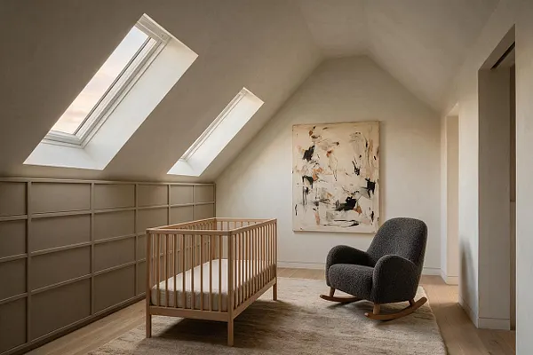 A modern nursery in a sloped-ceiling attic room with two skylights, a light wood crib, and a dark textured rocking chair on a cream rug. A large abstract painting and built-in wall paneling add a refined, minimalist feel.