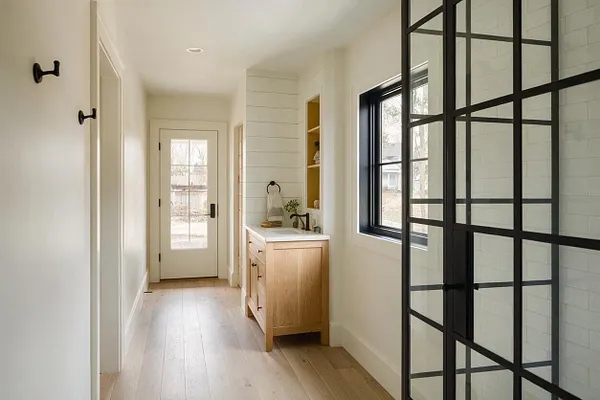 A bright farmhouse-style master bathroom with white shiplap walls, light wood flooring, and a natural wood vanity with a white countertop. A black-framed glass shower enclosure adds contrast on the right, while a window, minimal black hooks, and a glass-paned door bring in natural light.