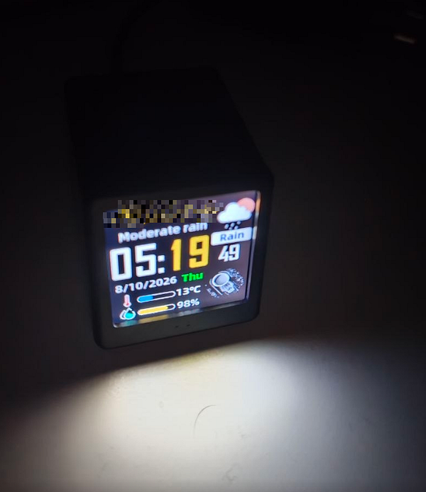 A digital alarm clock displaying the time 05:19 with weather and date information in a dimly lit room.