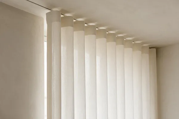 A minimalist interior detail showing tall cream vertical blinds or drapes hanging from a ceiling track beside a softly lit window. The room is finished in warm off-white tones with a clean, simple, monochromatic look.