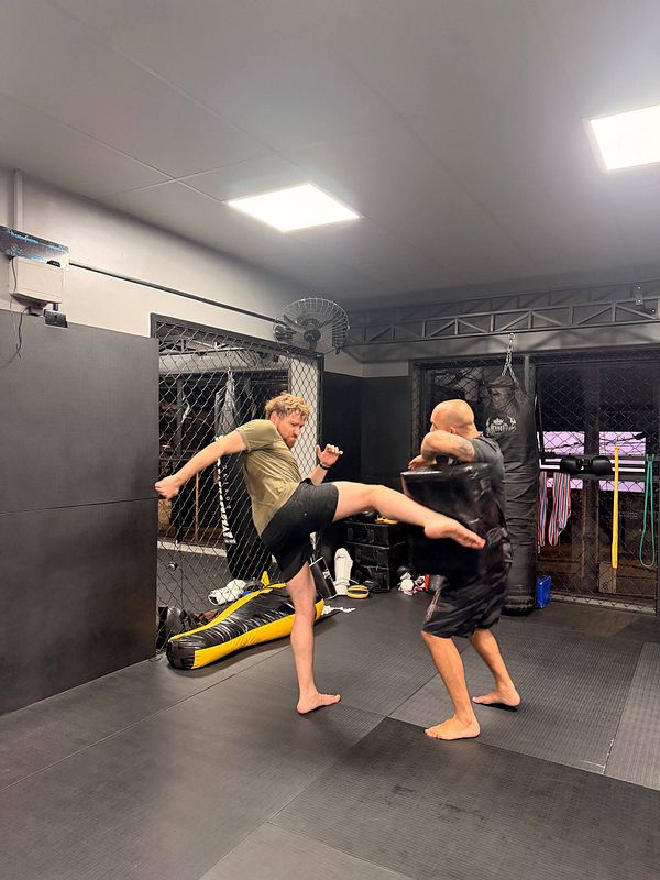 A male Muay Thai fighter is training in a gym, executing a high knee strike while wearing boxing gloves and shorts.