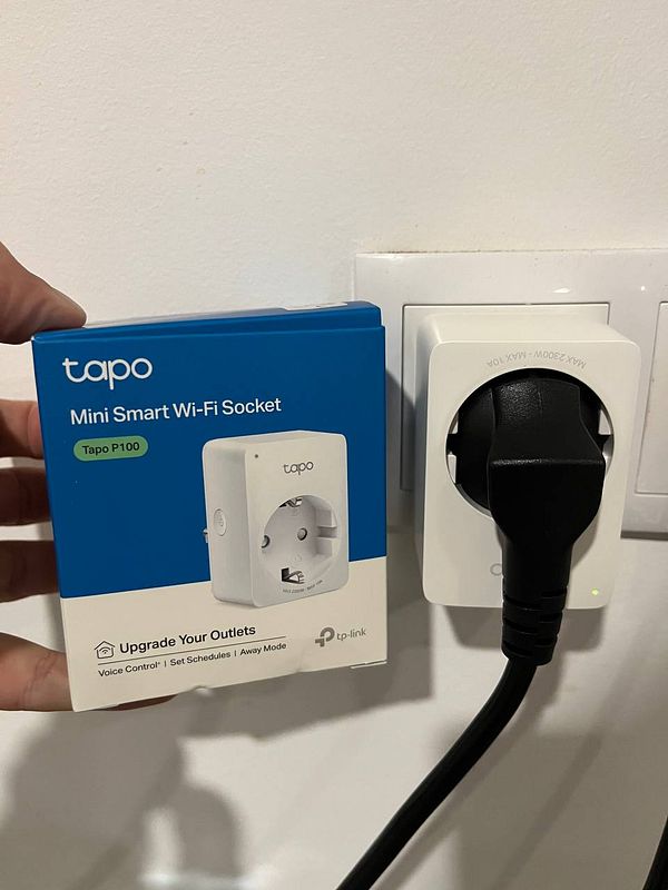 A hand holds a box for a Tapo Mini Smart Wi-Fi Socket next to the smart plug already installed in a wall outlet.