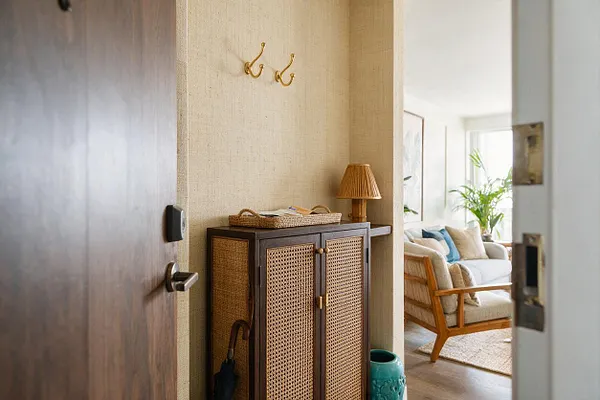 A tropical-style entryway seen through an open door, featuring a dark wood cabinet with woven rattan panels, a woven tray, and brass hooks on a textured beige wall. A small table lamp, umbrella, and turquoise vase add warmth, while a bright living room with light furniture and indoor plants is visible beyond.