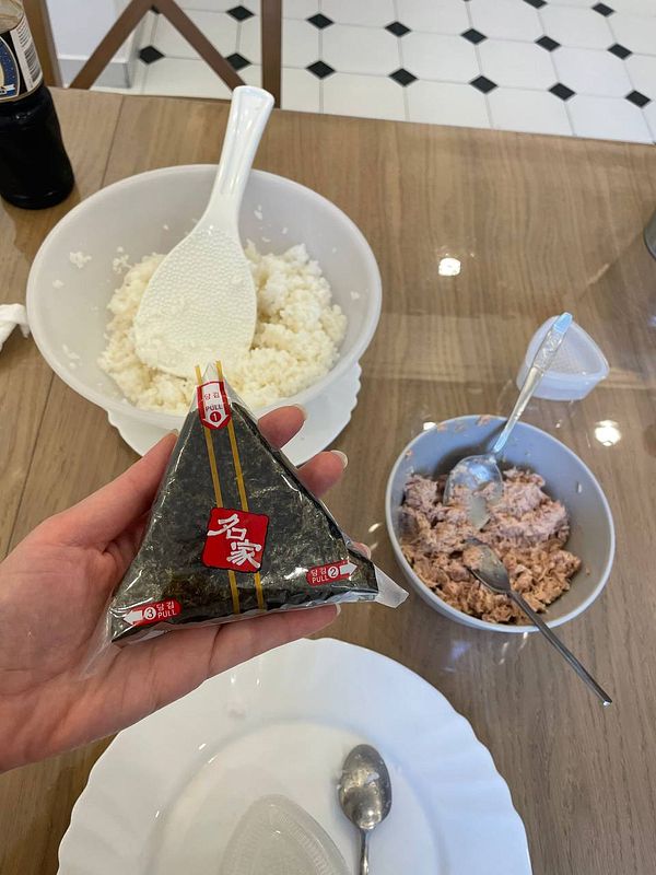 A hand holds a packaged onigiri in a kitchen setting with rice and tuna salad.