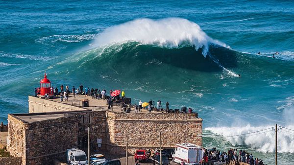 A massive wave crashes near a coastal lookout point in Nazaré, Portugal, with spectators gathered to watch.
