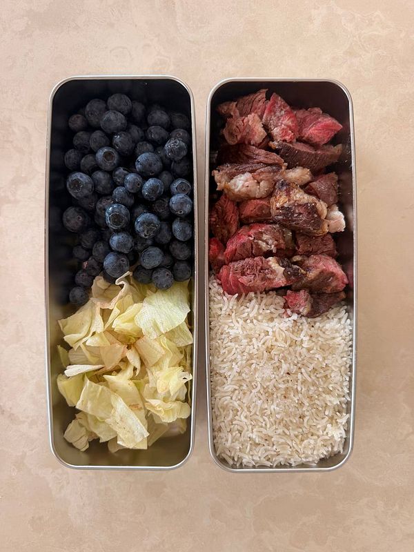 A packed meal consisting of steak, rice, fruit, and a beverage, presented on a white tray with utensils and a napkin.