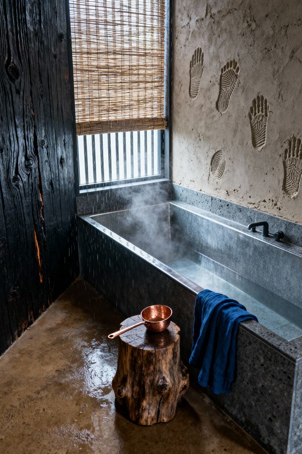 A Wabi-Sabi bathroom features a deep rectangular soaking tub in gray stone emitting steam beside a textured plaster wall marked with handprint impressions. A charred shou sugi ban wood wall and bamboo roll blind frame a window with black slats; a raw stump stool holds a copper ladle, an indigo towel drapes the tub edge, and matte black fixtures and a wet concrete floor reinforce the moody, elemental feel.