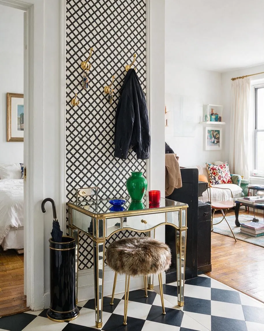 A bright studio apartment entry nook with a mirrored console table framed in brass, set against a black lattice wall panel. Gold hooks hold a dark coat above a fur-topped stool, and a black umbrella stand sits beside the table on a bold black-and-white checkered floor. The living area beyond includes white walls, colorful pillows, and warm wood flooring.