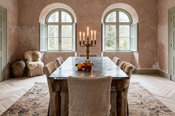 A French Country dining room features two arched windows with pale sage shutters lighting a long zinc-topped farmhouse table. Linen slipcovered chairs line the sides, with sheepskin throws on end chairs. Blush limewash plaster walls, light oak herringbone flooring, and a faded floral rug set a warm, timeworn tone. A brass candelabra with lit candles, peaches, and a jar of preserves sit at the center; an earthenware pot rests in the corner.