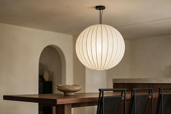 A modern dining area with a large spherical paper pendant light hanging above a dark wood table. Black dining chairs, a small sculptural bowl, and soft beige walls with an arched doorway create a calm, minimalist setting.