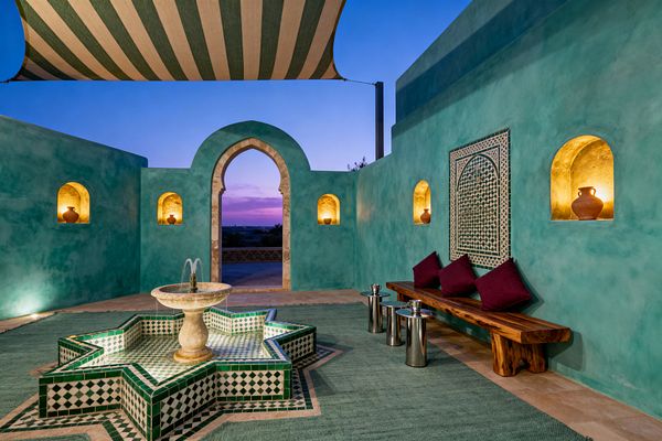 An open-air Moroccan patio at dusk with turquoise tadelakt walls and a limestone horseshoe arch, centered by an eight-pointed star fountain clad in green-and-white zellige and topped by a marble bowl. Gold-toned wall niches holding terracotta pots, a live-edge wooden bench with burgundy cushions, polished metal side tables, a sage flatweave rug, and a striped canvas canopy create a warm, intimate setting.