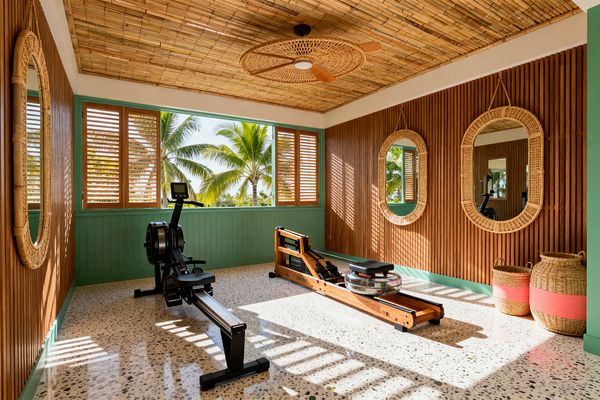 A bright home gym features a bamboo ceiling with a woven rattan fan, vertical wood-slat walls, and sea-green beadboard wainscoting. Wood louvered windows open to palm trees, casting striped light over a speckled terrazzo floor. A black rowing machine and a wood water rower face the windows, while three round rattan mirrors and two woven baskets with coral bands decorate the room.