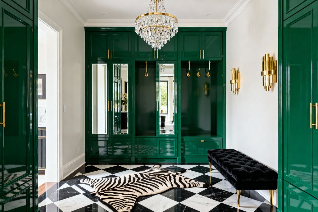 A glamorous mudroom showcases high-gloss emerald green cabinetry with mirrored door panels, polished brass pulls, and open cubbies fitted with brass coat hooks. A crystal chandelier and tiered brass wall sconces light a black-and-white marble checkerboard floor layered with a zebra-pattern hide, while a tufted black velvet bench with brass legs sits to the right; white walls and crown molding frame the space and a doorway reveals light from an adjacent room.