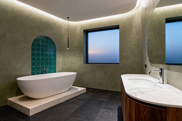 A contemporary bathroom with a matte white freestanding tub on a light travertine plinth, set before an arched alcove clad in teal patterned tiles and fitted with wall-mounted chrome fixtures. Curved sage plaster walls flow into a coved ceiling illuminated by concealed LED lighting, with a slim black pendant above the tub. To the right, a rounded walnut vanity with a white marble countertop stands on charcoal large-format floor tiles beside a frosted window.