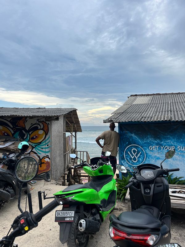 A person stands on the beach, gazing at the ocean under a cloudy sky, with parked scooters in the foreground.