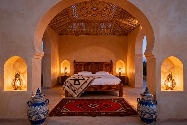 A Moroccan-style bedroom framed by stucco arches features a carved wooden bed beneath a pitched reed ceiling, with warm brass lanterns glowing inside arched wall niches and on bedside tables, casting amber light on sand-colored polished plaster. A red diamond-pattern Berber rug grounds the space while a black-and-cream geometric throw tops the white bedding, and two large blue-and-white ceramic urns sit near the entry.