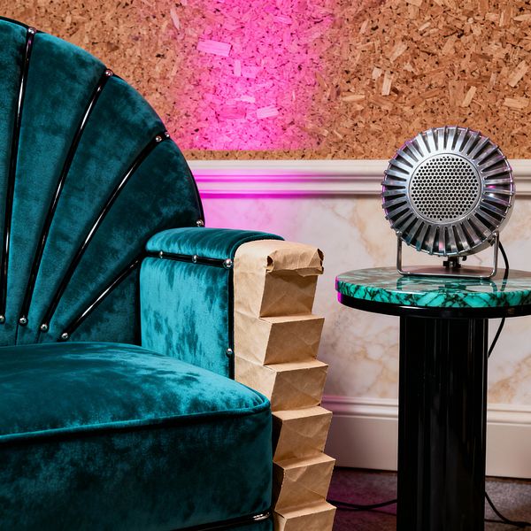 Close-up of an Art Deco media room corner with a scalloped, fan-back armchair upholstered in teal velvet, edged with black piping and small metal studs, and a stepped insert along the outer arm. Next to it is a round side table with a green malachite top on a glossy black pedestal, holding a chrome radial microphone-style speaker. Behind, cork wall panels glow magenta above cream marble wainscoting with white trim, over dark carpet with a visible cable.
