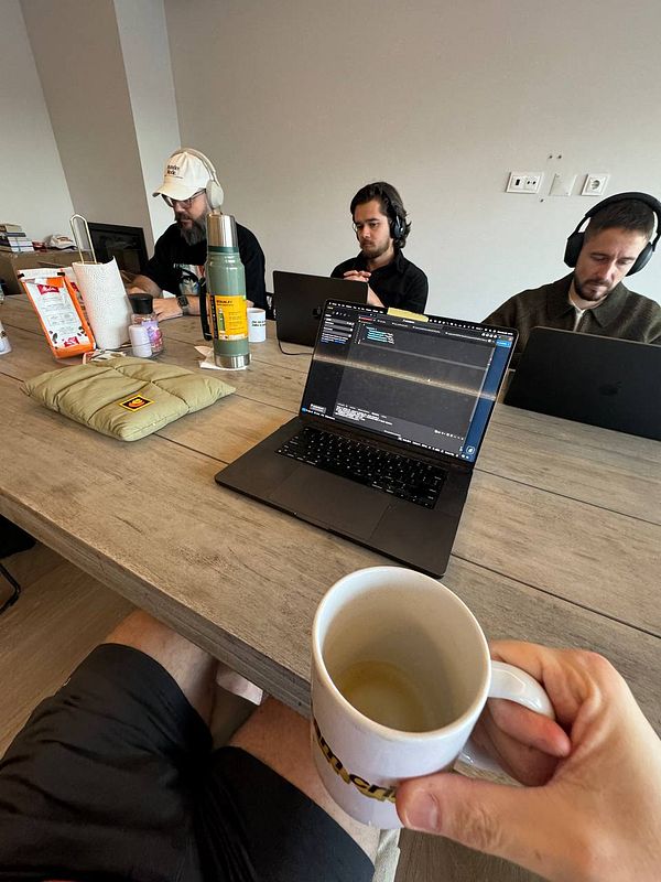 A group of three individuals is engaged in a coworking session at a table, with laptops and personal items around them.