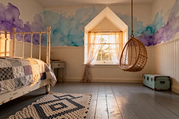 A sunlit children’s bedroom features a distressed white iron bed with a patchwork quilt, a rattan hanging chair suspended by rope, and a gabled window dressed in sheer curtains. Beadboard wainscoting lines the lower walls beneath a lavender and aqua watercolor mural, with a weathered bedside table, vintage turquoise trunk, gray-painted plank floors, and a chunky crocheted rug completing the space.