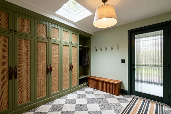 Transitional mudroom with olive built-in lockers featuring cane inserts and leather strap pulls, plus an open cubby with hanging coats and shoe storage. A sculpted walnut bench sits below a row of metal hooks opposite a deep green door with reeded glass, while a skylight and linen drum pendant illuminate checkerboard terrazzo flooring and a striped rug against soft sage walls.