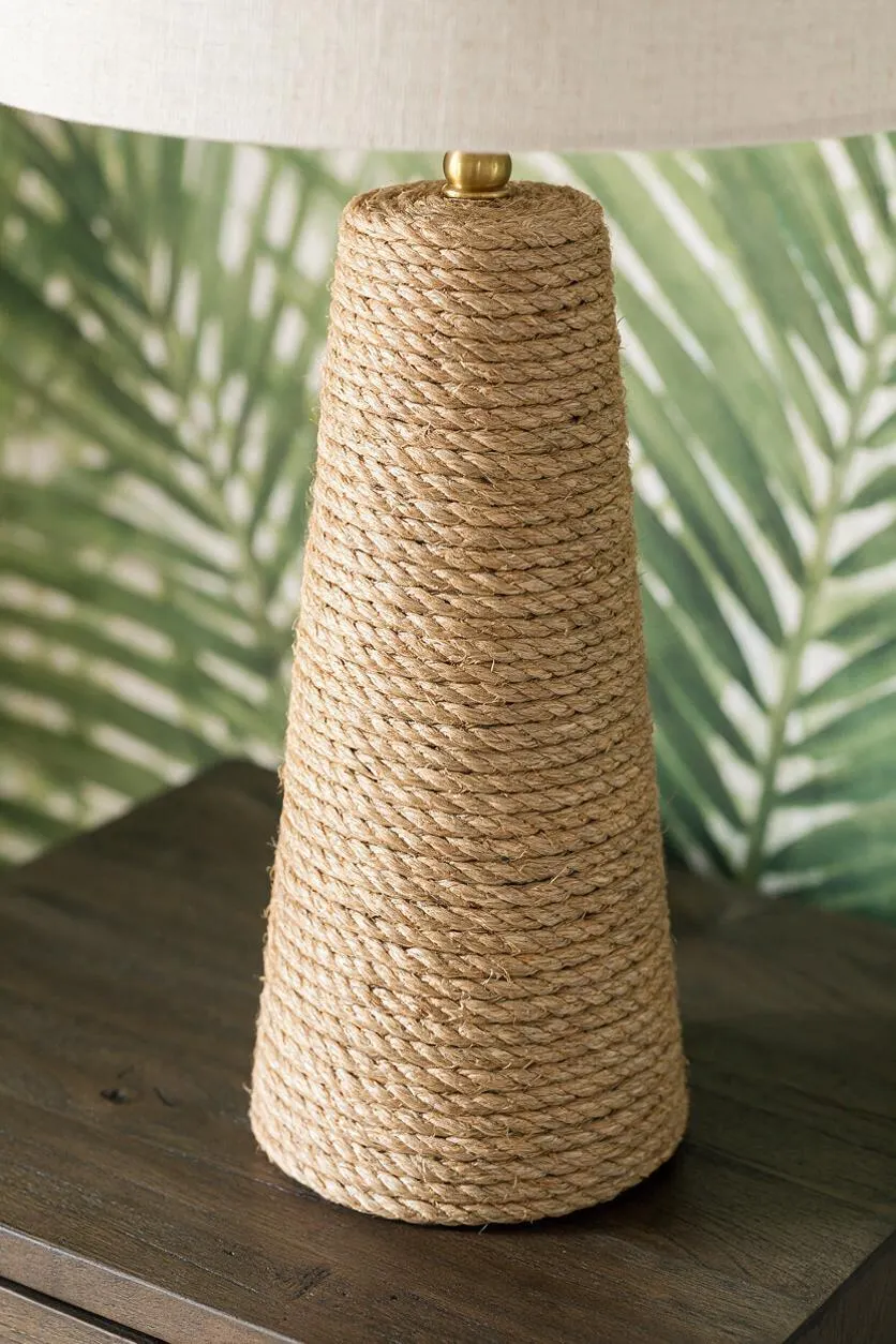 A table lamp with a conical base tightly wrapped in natural rope sits on a dark wood surface. The lamp has a light fabric shade and a small brass accent at the top, with a blurred tropical palm-leaf background behind it.