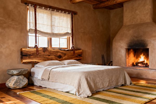 A rustic bedroom with clay-colored plaster walls features a low bed in beige linen, anchored by a thick live-edge wood headboard suspended with leather straps beneath gauzy linen window shades. To the right, a wood-burning fireplace with a sculpted plaster surround glows, while exposed ceiling beams, wide-plank wood floors, a stacked-stone bedside table, and a mustard-and-olive striped rug add natural texture and a warm, earthy palette.
