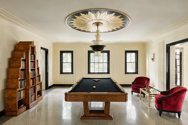 A bright Art Deco game room centered on a wood pool table with a black felt top, flanked by two red velvet chairs and a gold glass side table. The room features cream walls with vertical detailing, black-trimmed windows and doorways, a built-in geometric wood bookcase, and a dramatic starburst ceiling medallion with a hanging chandelier.