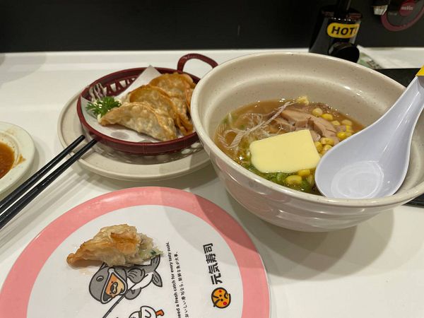 A table setting featuring a half-eaten fried dumpling on a sushi plate, a basket of additional dumplings, and a bowl of ramen with corn, chicken, and a slab of butter with a white soup spoon.