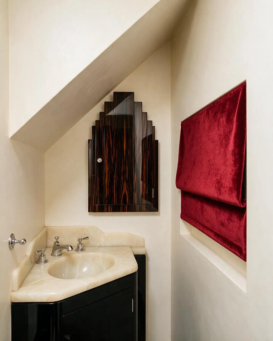 A compact powder room with a sloped ceiling features a cream marble vanity, a glossy dark wood Art Deco cabinet above the sink, and deep red velvet window shades on the right wall. The palette pairs soft ivory walls with black, burgundy, and warm stone tones for a dramatic, elegant look.