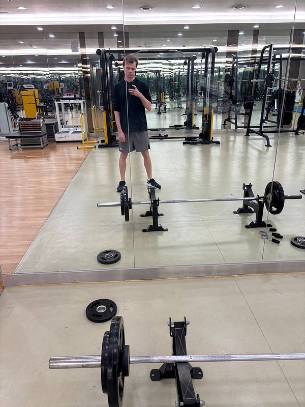A person stands in a gym, taking a selfie in front of a mirror with gym equipment in the background.