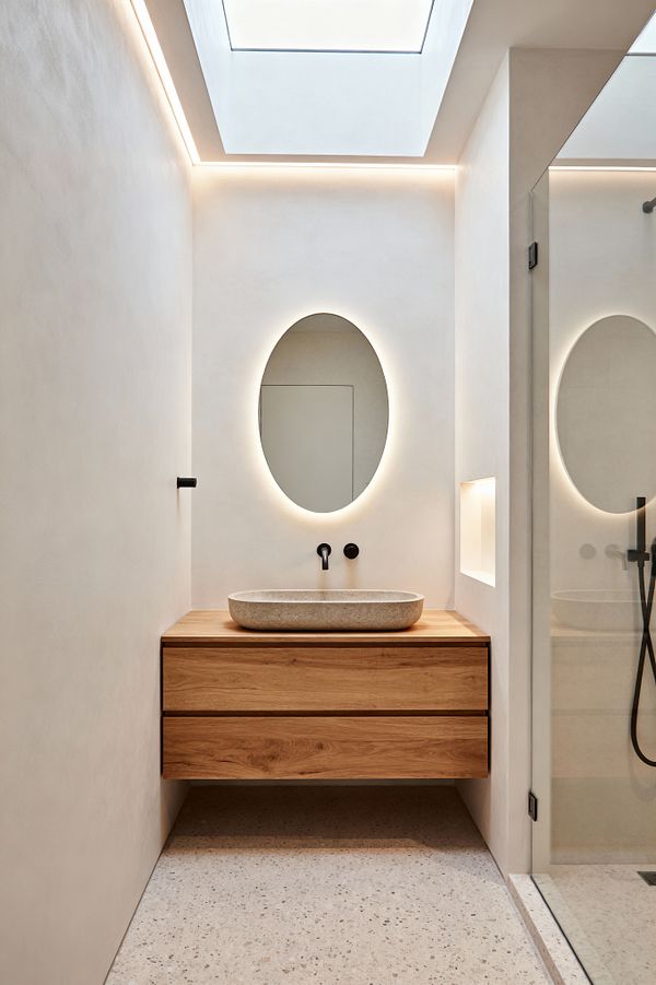 A narrow minimalist bathroom with cream plaster-like walls features a floating oak vanity and a shallow beige stone vessel sink. An oval mirror with halo LED backlighting sits beneath a large skylight and subtle cove lighting; to the right is a clear-glass shower with matte black fixtures and a small recessed niche, all set on light speckled terrazzo flooring for a calm, uncluttered look.
