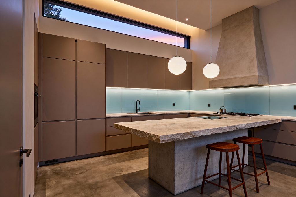 A contemporary kitchen features matte taupe, handleless cabinets in a U shape around a large island topped with a rough-cut stone slab on a concrete base. A pale aqua back-painted glass backsplash runs the perimeter with undercabinet LEDs; a sculptural concrete range hood anchors a gas hob, a flush black cooktop sits in the island, and two opal globe pendants, a clerestory window, and cove lighting illuminate polished concrete flooring and two reddish wood stools.
