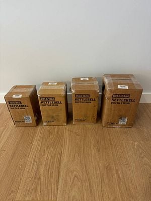 Four boxes of kettlebells are lined up on a wooden floor against a plain wall.