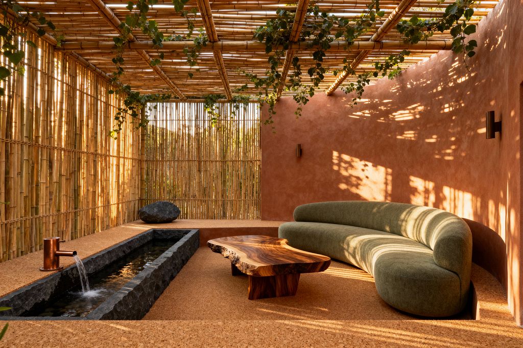 A biophilic living room is enclosed by vertical bamboo screens and an overhead bamboo trellis threaded with trailing vines, which cast dappled shadows on a terracotta plaster wall and cork floor. An olive green curved sofa faces a live-edge wood coffee table. Alongside, a narrow basalt water rill with a copper spout flows gently beside a large dark stone, creating an open-air, serene atmosphere.