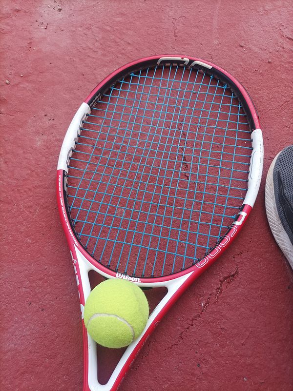 A tennis racket and a tennis ball are resting on a red court surface.