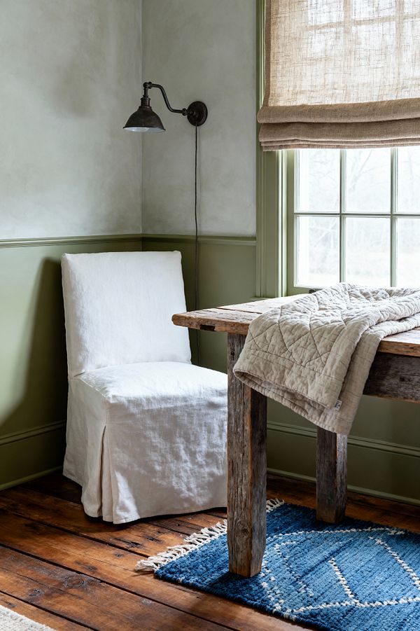 Farmhouse living room corner featuring a rustic reclaimed-wood table by a window with sage green trim and a natural linen Roman shade. A white linen slipcovered chair sits on wide-plank pine floors with a small indigo rug; an oatmeal quilt is draped over the tabletop, and a vintage blackened-iron wall sconce hangs above pale plaster walls, all softly lit by daylight.