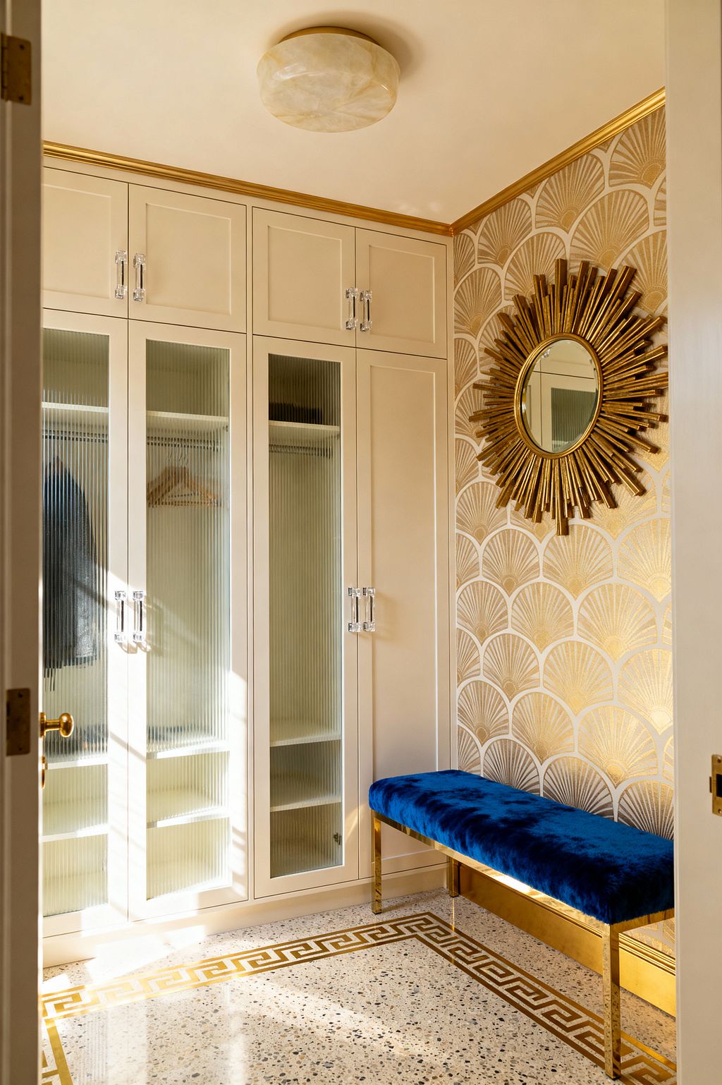 A glamorous mudroom features tall cream cabinetry with reeded-glass doors and clear acrylic pulls, while gold fan-pattern wallpaper holds a gilded sunburst mirror above a royal blue velvet bench with a brass base. A terrazzo floor framed by a gold Greek key inlay, an alabaster flush-mount light, and gold crown molding add warm illumination, with open cubbies and a hanging rail visible inside the cabinets.
