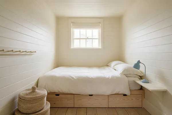 A narrow coastal guest bedroom with white shiplap walls, a small centered window, and a low platform bed dressed in white bedding. Light wood storage drawers sit beneath the bed, with a blue bedside lamp on a floating shelf and woven baskets adding natural texture.