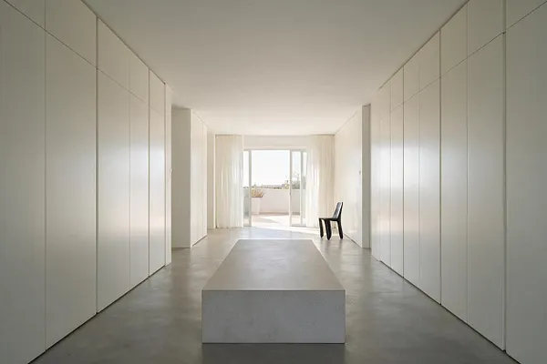 A long minimalist closet with floor-to-ceiling white cabinet doors lining both sides of a narrow room. A simple rectangular bench sits in the center, with a black chair near bright sliding doors and sheer curtains at the far end.