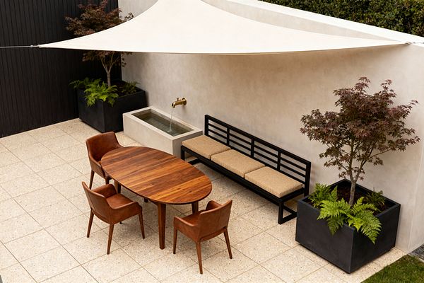 An outdoor patio with a cream shade sail covers a space arranged with an oval walnut dining table and four cognac leather chairs, opposite a black metal bench with three tan cushions along a warm off-white plaster wall. A brass spout pours into a rectangular concrete fountain, large black planters hold a Japanese maple and ferns, and speckled cream terrazzo pavers set a clean grid, with a black slat fence at the left.