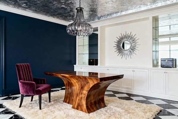 Home office with a sculptural, high-gloss walnut desk centered on a cream shag rug over black-and-white marble tiles. To the left sits a plum velvet, channel-tufted chair against a deep navy wall; to the right, white built-in cabinetry with brass knobs, mirrored shelves, and a silver sunburst mirror. A smoky glass chandelier hangs from a silver-leaf ceiling, reflecting daylight and adding glamorous sparkle throughout the room.
