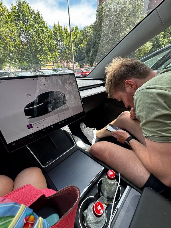 A person is seated in a car, focused on their phone while a large touchscreen display shows a vehicle interface.