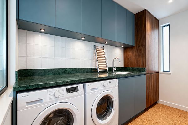 A contemporary laundry room features matte blue-gray handleless cabinets above and below a deep green stone countertop, with a glossy white square-tile backsplash illuminated by recessed under-cabinet spotlights. A white undermount sink with a stainless steel faucet sits beside front-loading washer and dryer units, while a tall walnut cabinet, slim black-trim window, cork flooring, and a compact leaning drying rack complete the clean, functional space.