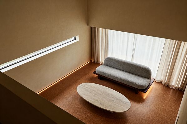 A minimalist living room seen from above features a low gray bouclé sofa on a dark wood base set beside full-height windows with beige sheer curtains. An oval light-stone coffee table sits on warm cork flooring. A slim horizontal window breaks up a smooth beige wall. Concealed LED strips run along the perimeter and beneath the sofa platform, casting a soft warm glow that emphasizes clean lines, negative space, and an uncluttered layout.
