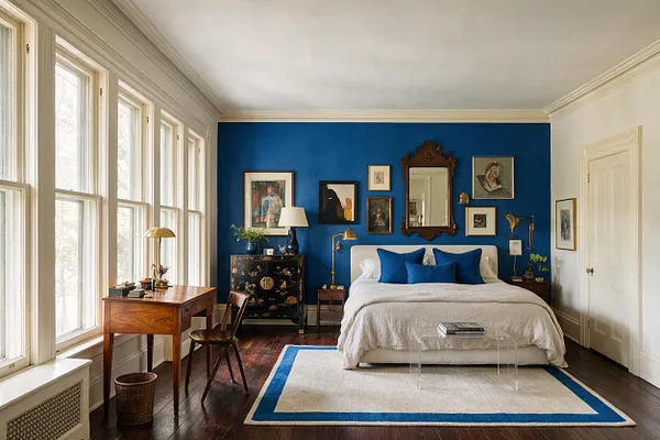 A guest bedroom with a vivid cobalt blue accent wall behind a white bed, layered with framed artwork and a carved wood mirror. Large tall windows line the left side, while a wooden desk, vintage chair, brass lamps, and a clear acrylic coffee table add eclectic character over dark wood floors and a cream-and-blue rug.