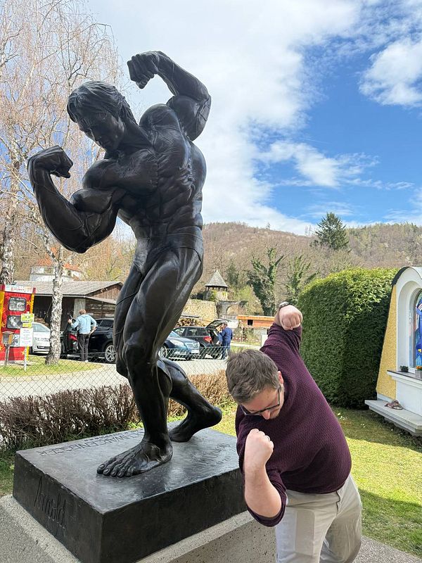 A smiling man wearing a gray beanie and dark jacket stands next to a colorful dinosaur and a large blue arrow sign outside a building.