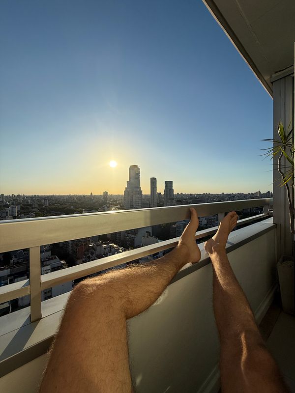 A person relaxes on a balcony with their legs stretched out, overlooking a city skyline at sunset.