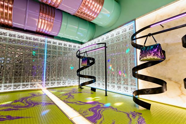 An exuberant room with glass-block walls lit from behind, a creamy backlit stone panel, and exposed mint and lavender ceiling ducts wrapped with copper bands. The floor is acid chartreuse rubber with purple marbling and an illuminated strip through the center. RGB LEDs trace edges, casting multicolor reflections. Sculptural black steel spirals and a minimal frame stand hold iridescent sequined bags, creating a glossy, neon-infused environment.
